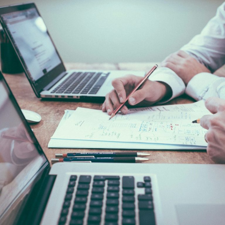 Hände, die Notizen machen, umgeben von zwei geöffneten Laptops und Stiften.