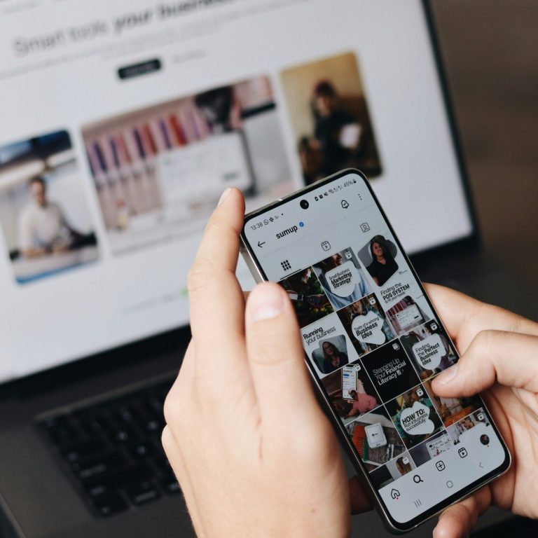 Eine Person hält ein Smartphone mit Social-Media-Inhalten vor einem Laptop.