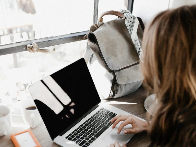 Eine Person arbeitet an einem Laptop, im Hintergrund eine Tasche und ein Tisch mit Getränk.