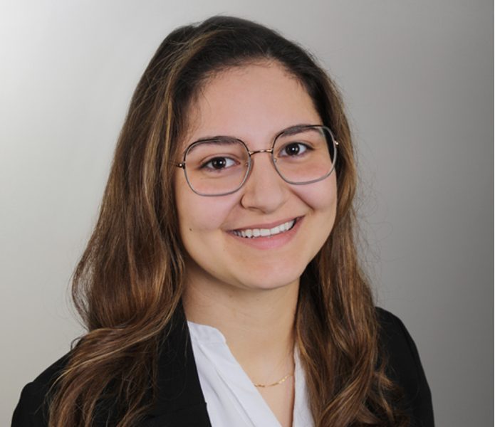 Junge Frau mit Brille, lächelnd, trägt einen schwarzen Blazer und ein weißes Oberteil.