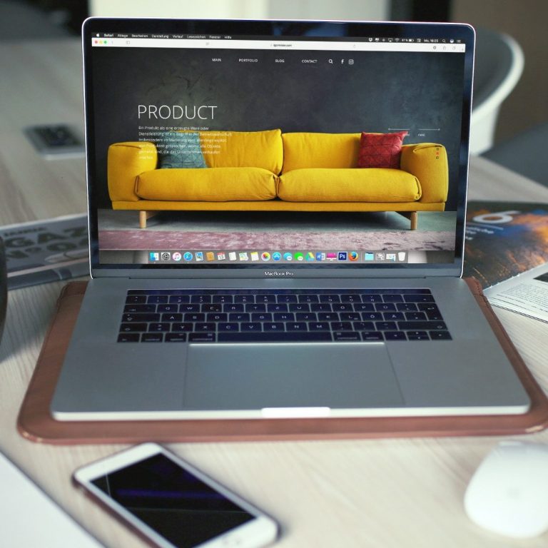 Laptop mit gelbem Sofa auf dem Bildschirm, daneben Smartphone und Unterlagen.